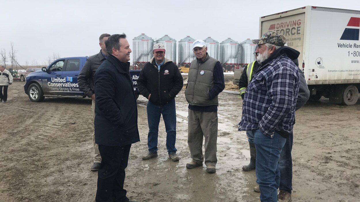 Jason Kenney entouré d'un groupe de fermiers. Il sourit. À l'arrière-plan, il y a des silots de grain.