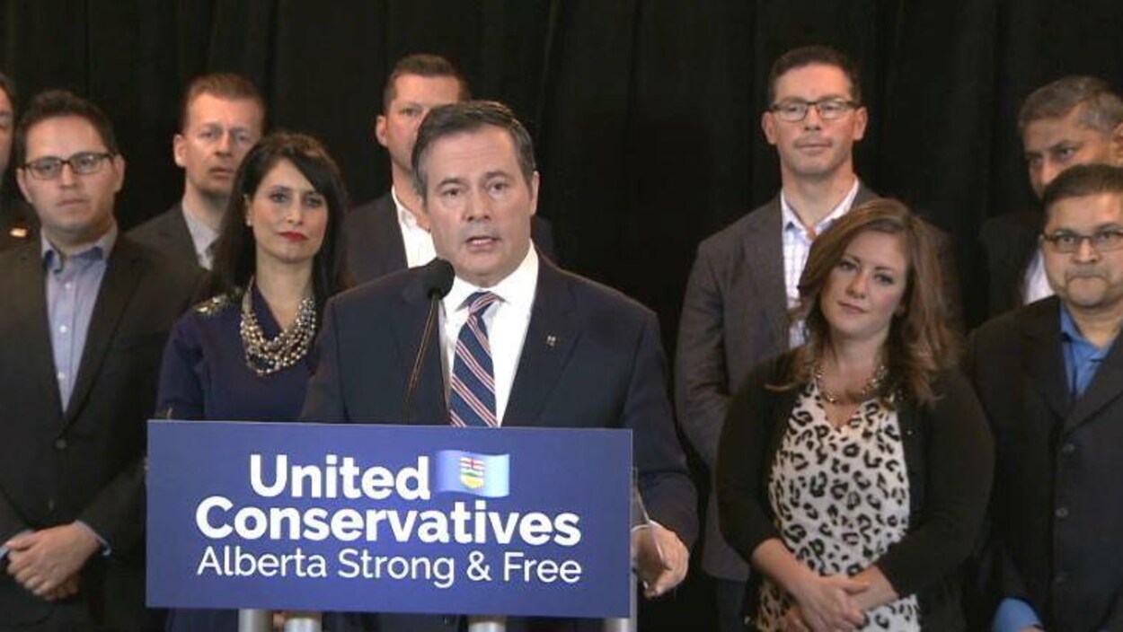 Jason Kenney se tient derrière un lutrin, entouré de candidats de son parti.