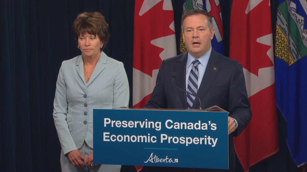 Jason Kenney parle devant un podium. La ministre se tient debout à sa droite.