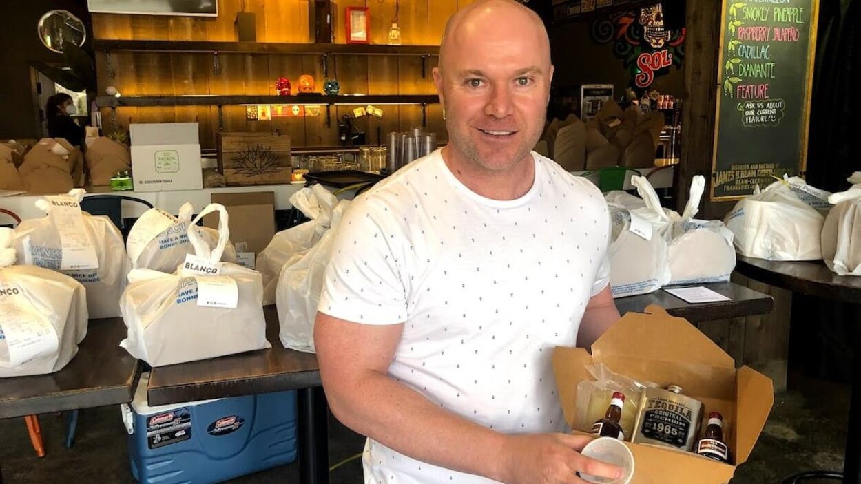 Un homme chauve tient une boîte en carton avec différent type d'alcools pour préparer une margarita chez soi.