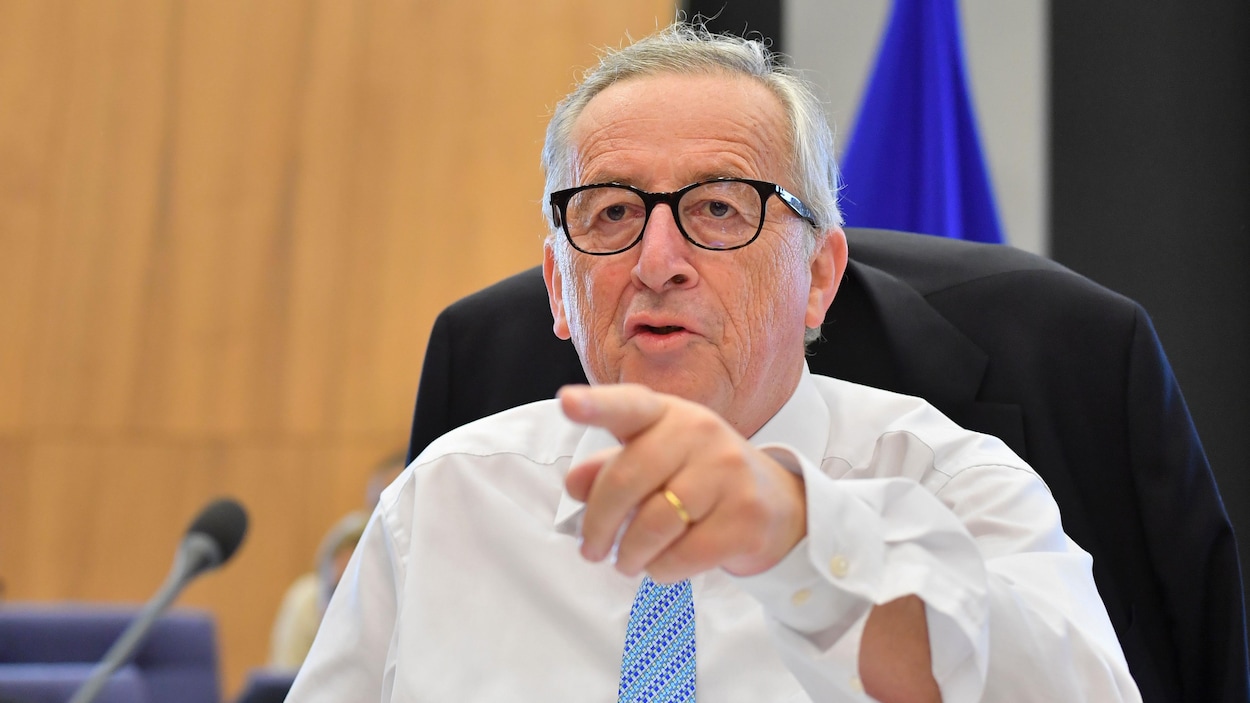 Un homme portant une blouse et une cravate montre du doigt devant lui avec son index gauche. L'homme porte des lunettes et a les cheveux gris. Un homme portant une blouse et une cravate montre du doigt devant lui avec son index gauche. L'homme porte des lunettes et a les cheveux gris.