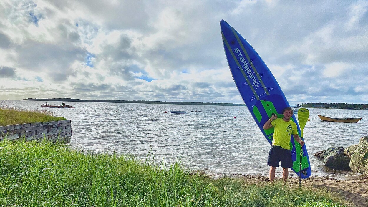 Un homme tient une planche à pagaie devant une plage.