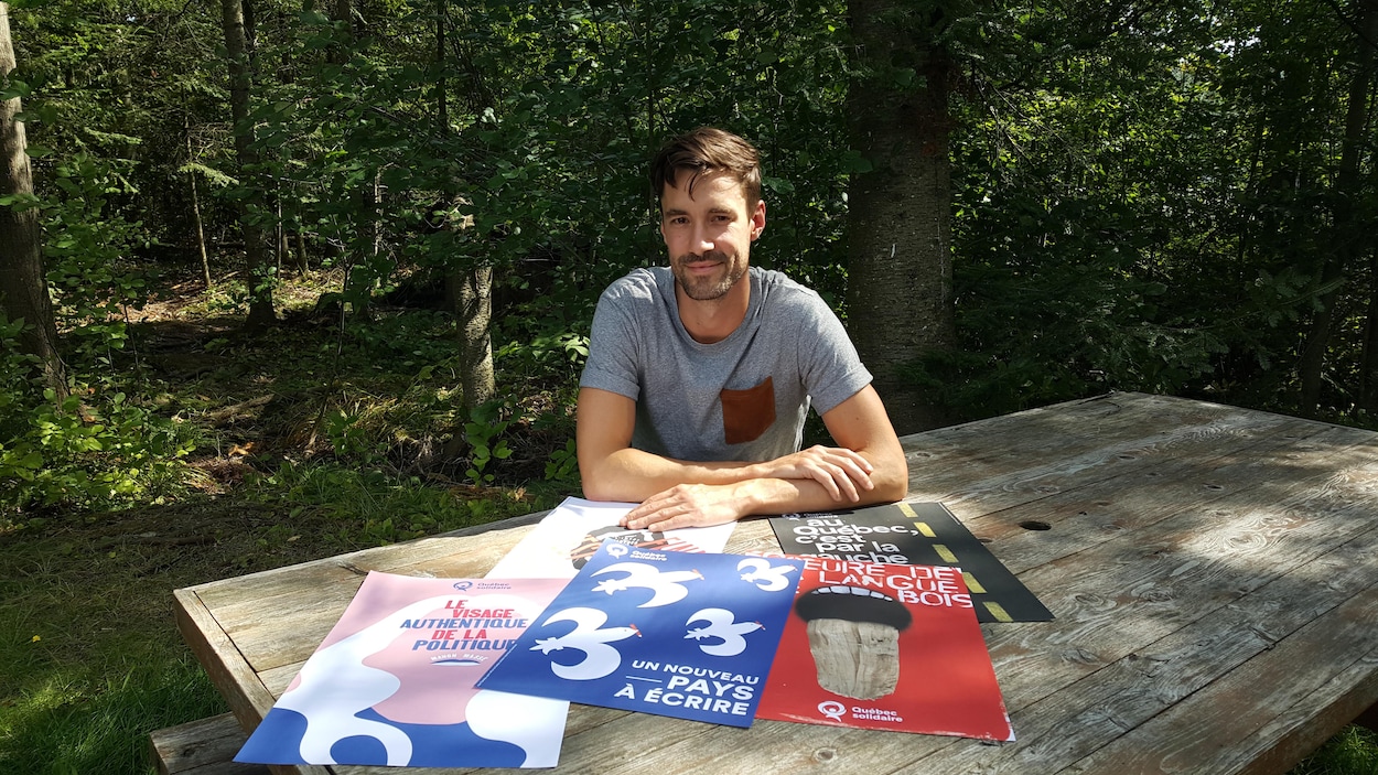 Une jeune homme d'une trentaine d'années est assis à une table à pique-nique. Sur la table sont exposés cinq affiches électorales.