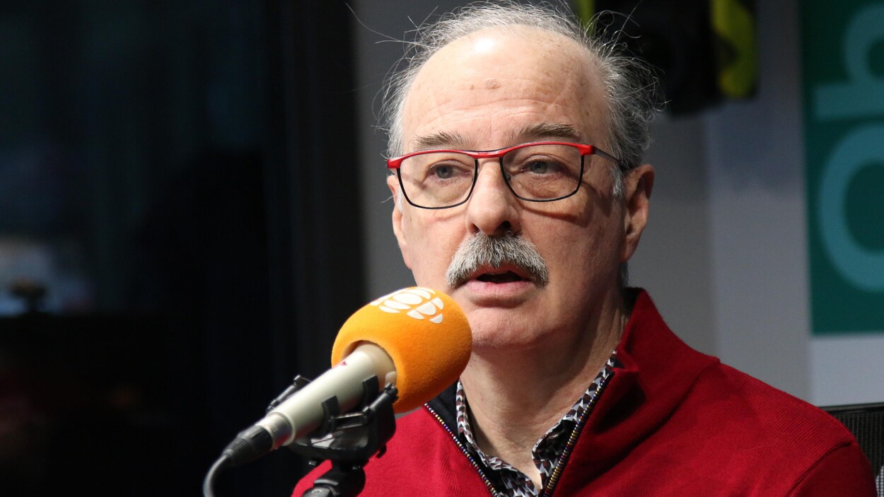 Un homme avec une moustache et des lunettes derrière un micro dans un studio de radio.