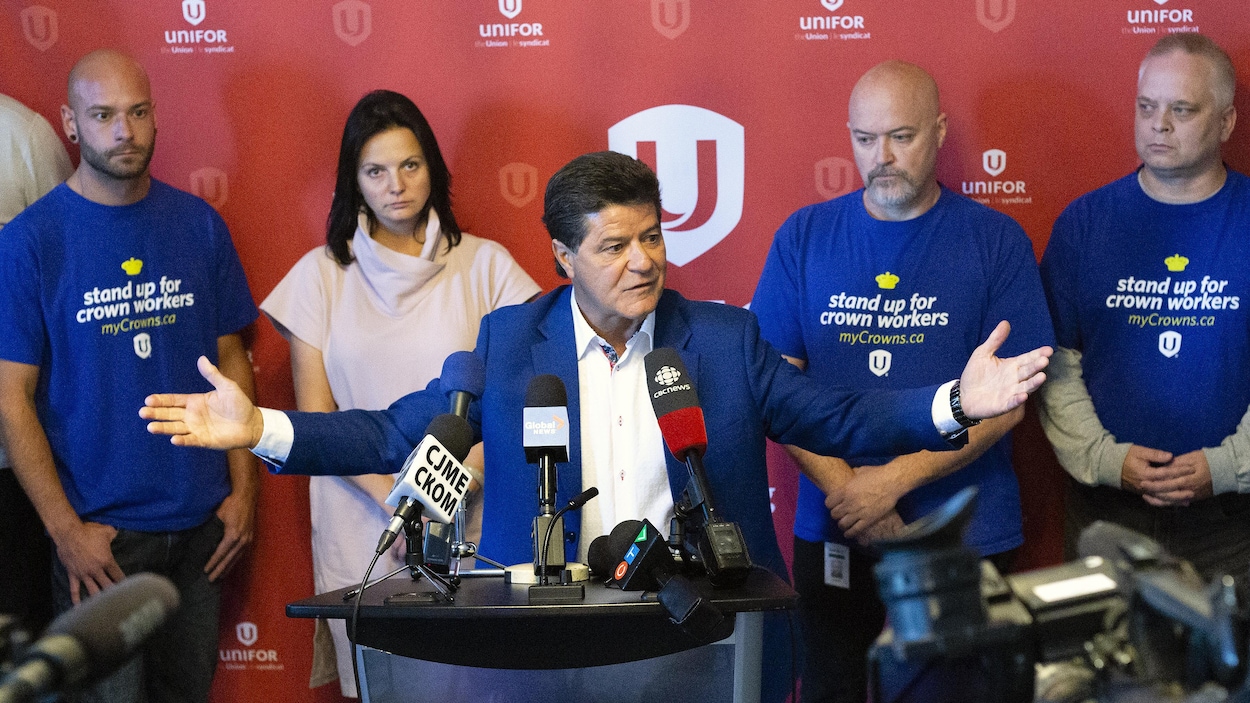 Jerry Dias a les bras ouverts devant les micros des journalistes. Plusieurs personnes se tiennent debout derrière lui.