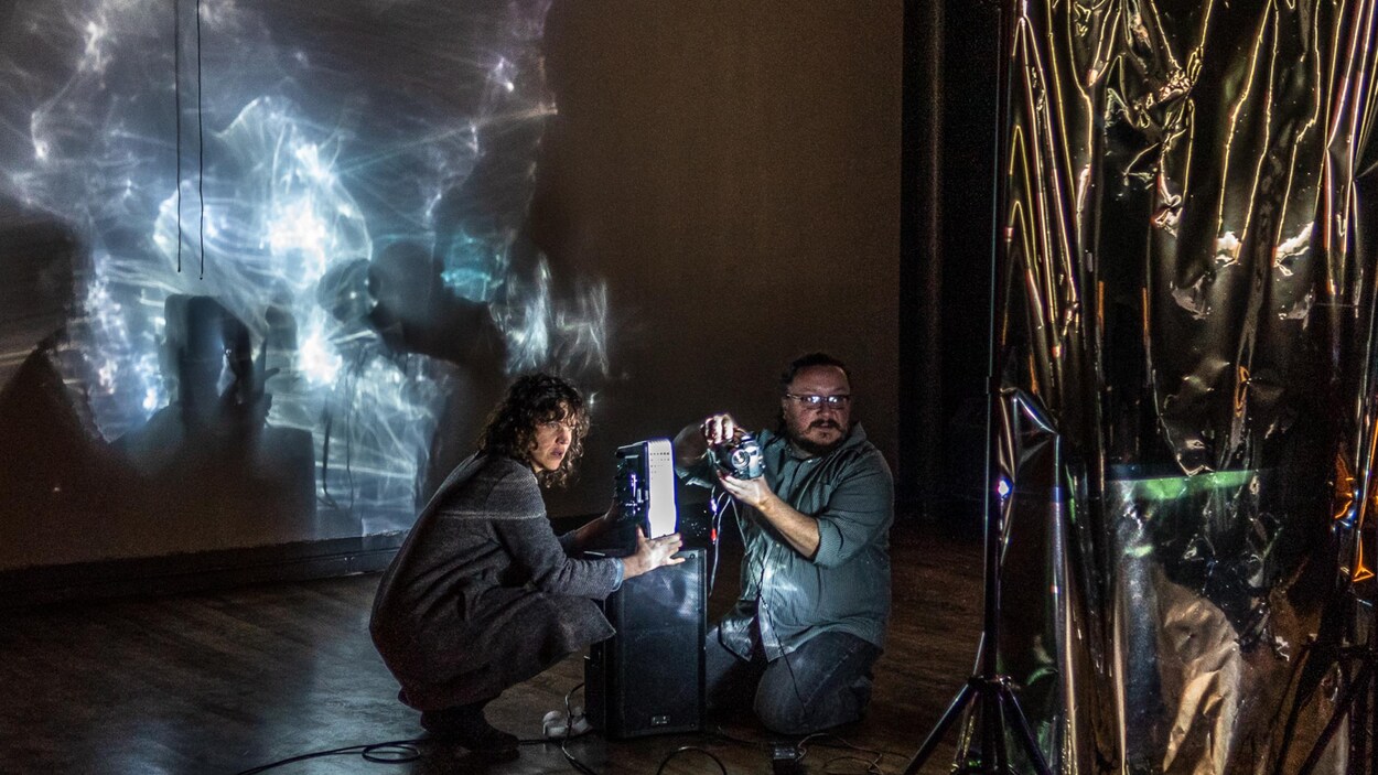 Les deux artistes sont accroupis près d'un projecteur et regardent une toile en plastique transparent.