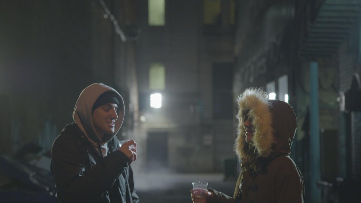 On voit deux jeunes personnes dans une ruelle, le soir, en train de consommer une boisson dans des verres en plastique.