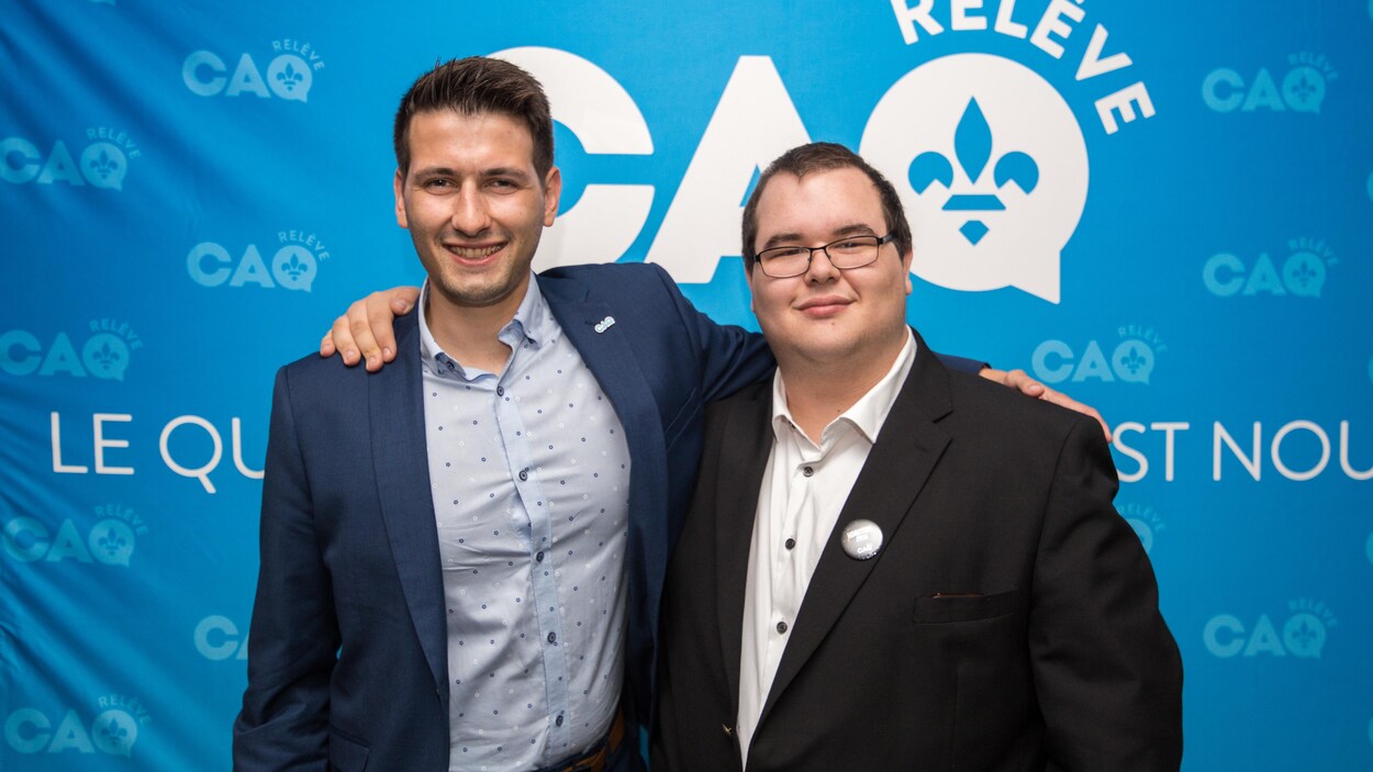 Deux hommes sourient à la caméra devant des logos de la relève de la CAQ.