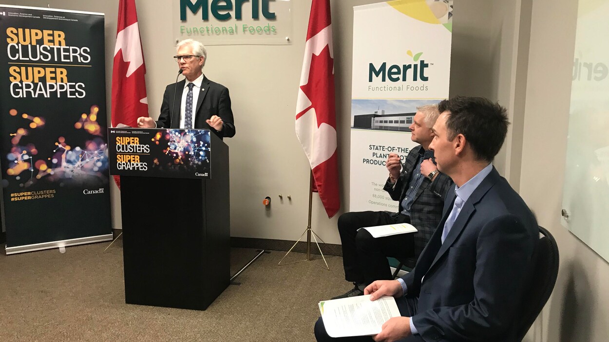 Jim Carr est en conférence de presse, devant deux drapeaux du Canada et une affiche de la compagnie Merit Functional Foods. Deux personnes sont assises à côté et l'écoutent.