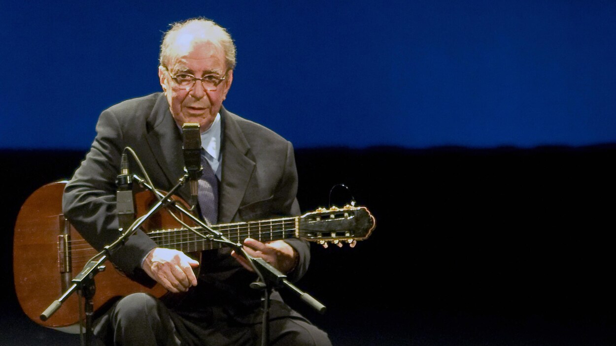 Le chanteur est assis face au micro, une guitare à la main, sur scène.