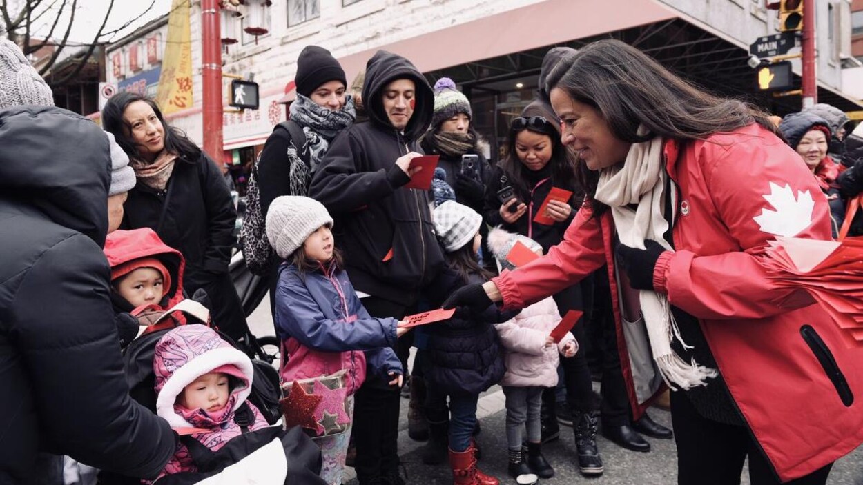 Une femme dans la quarantaine en veste rouge avec une feuille d'érable donne des feuilles rouges à des enfants et des adultes dans une rue lors d'un défilé.