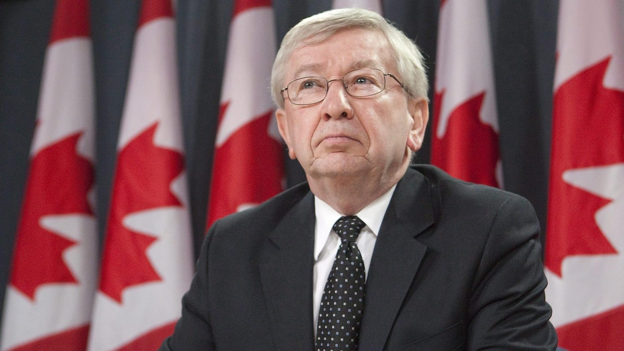 Un homme avec des lunettes, un veston et une cravate devant des drapeaux du Canada. Un homme avec des lunettes, un veston et une cravate devant des drapeaux du Canada.