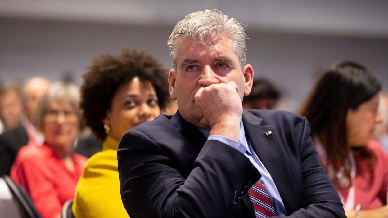 John fraser dans une assemblée assis écoute avec sa main contre son visage.