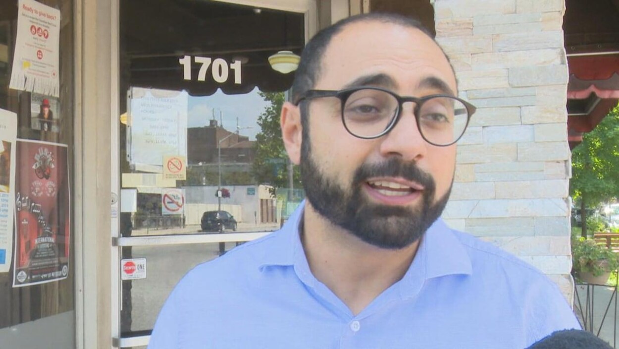Un homme avec des lunettes et une barbe devant un magasin répond à un journaliste