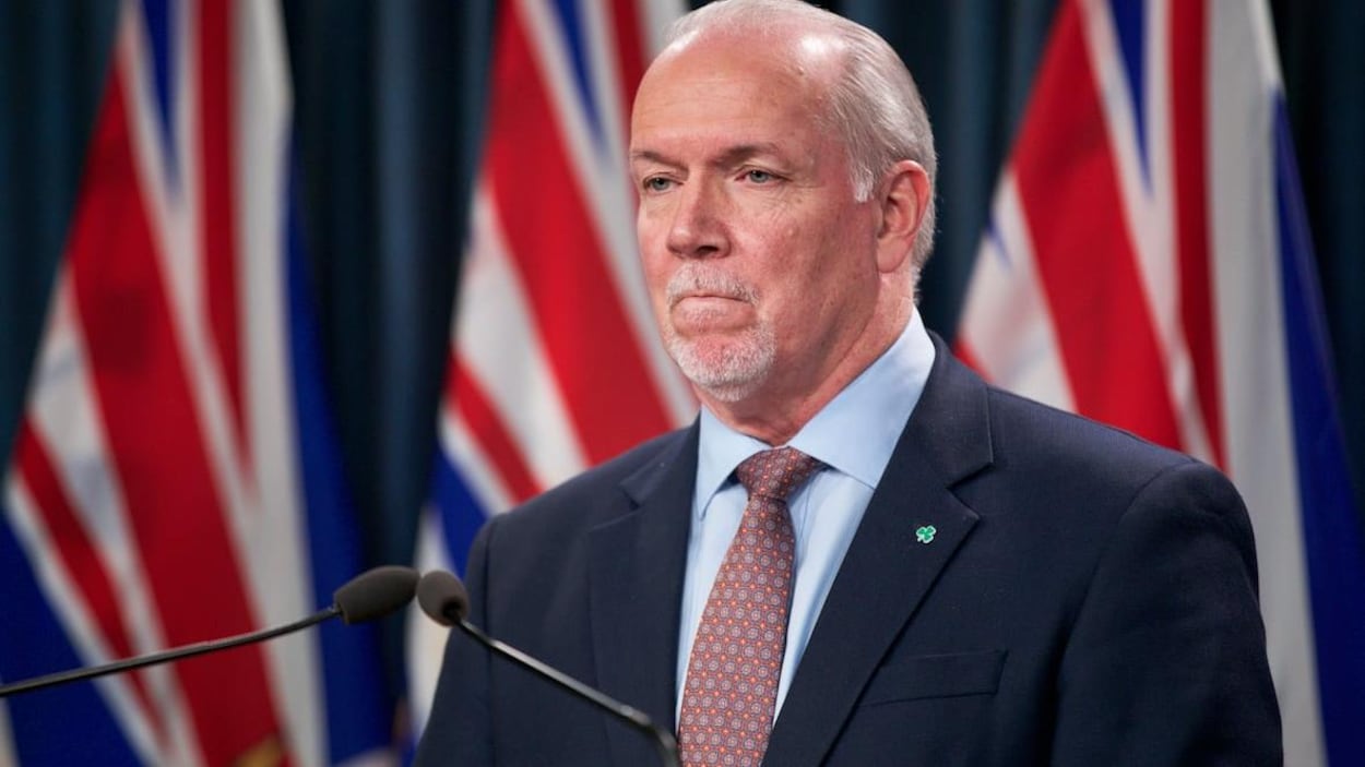 Le premier ministre de la Colombie-Britannique, John Horgan, en conférence de presse devant des drapeaux provinciaux.