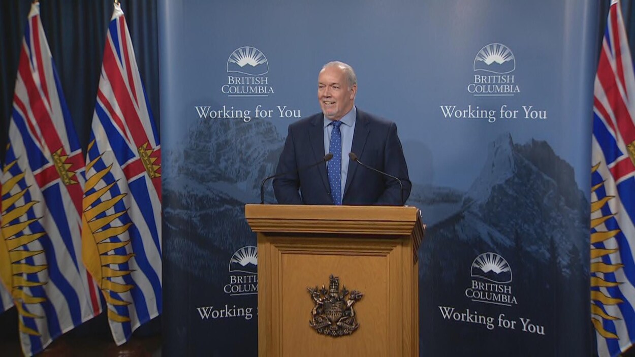 John Horgan est debout derrière un pupitre et s'adresse aux journalistes. On peut voir une affiche derrière lui avec le slogan écrit en anglais : La Colombie-Britannique à l'oeuvre pour vous.