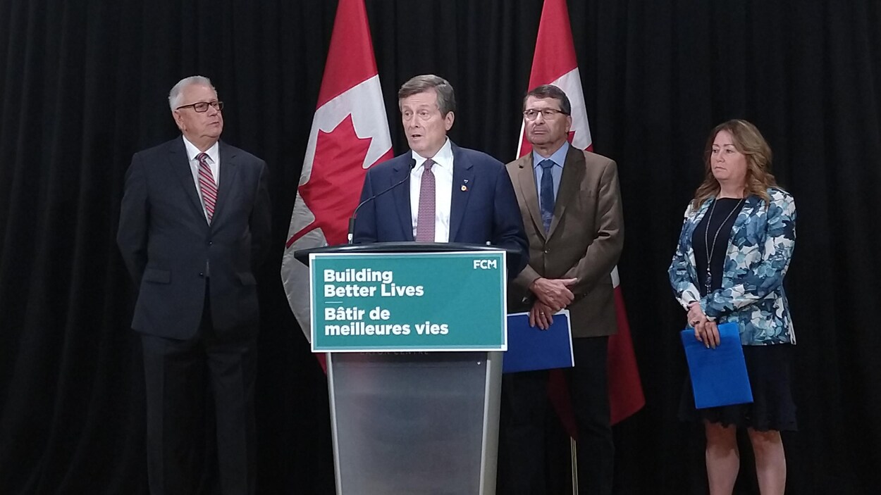 Quatre personnes sont derrière un podium, devant un rideau noir et deux drapeaux canadiens.