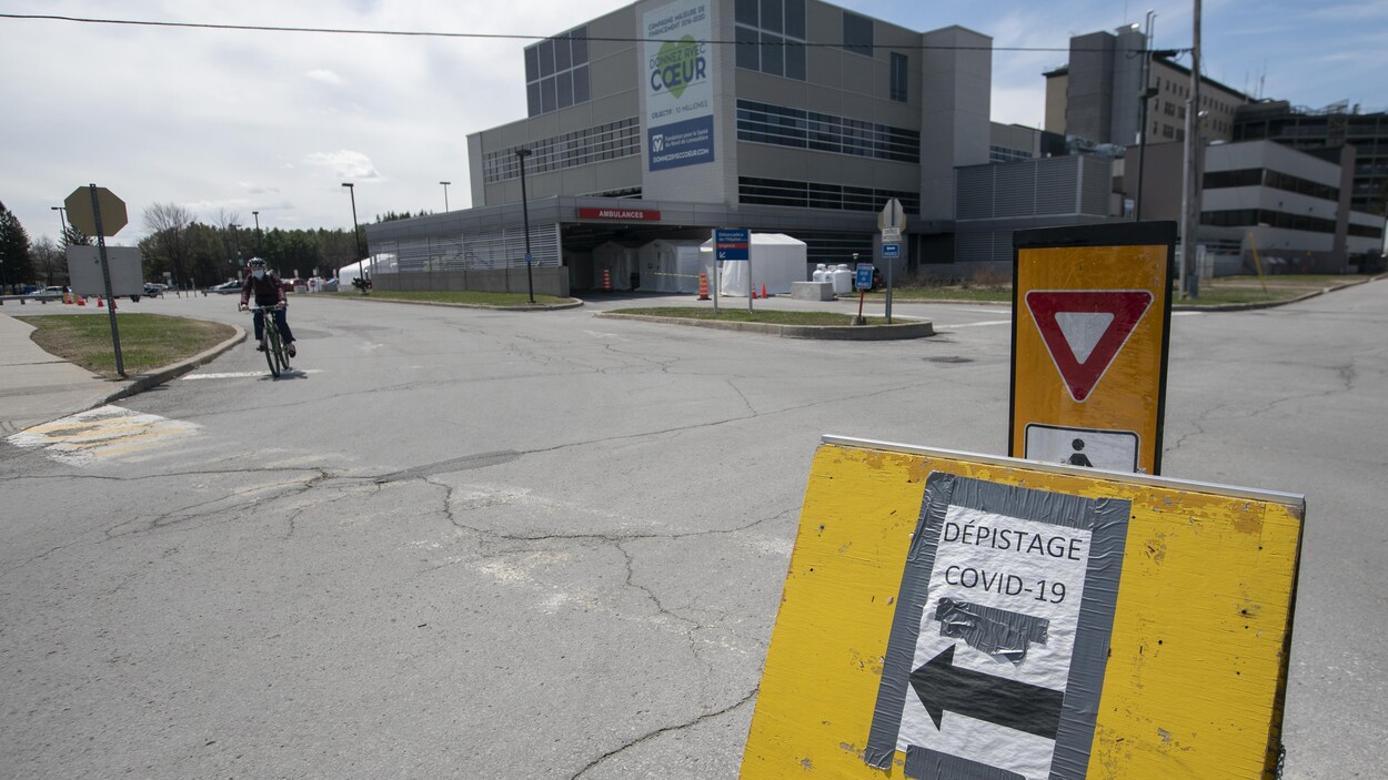 Un hôpital en arrière-plan ainsi qu'un panneau de bois sur lequel on peut lire « Dépistage COVID-19 ».