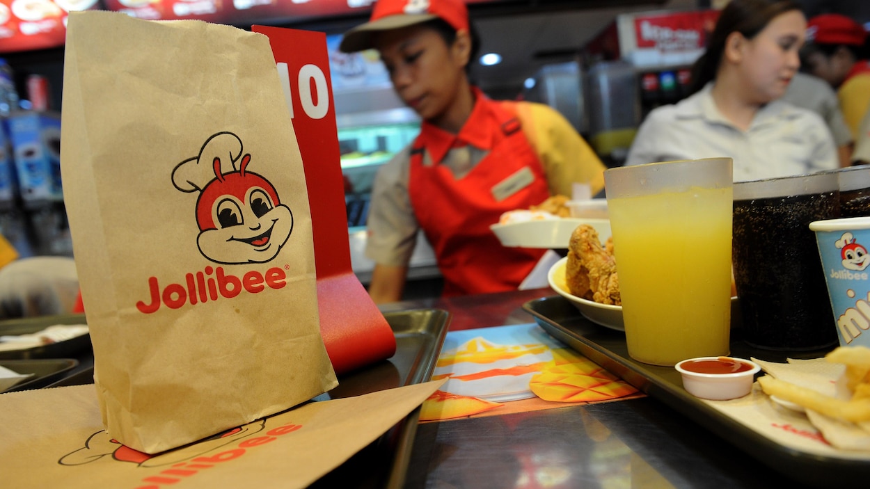 Deux plateaux contenant de la nourriture et des boissons servies par Jollibee avec, en arrière-plan, les employés qui préparent la nourriture.