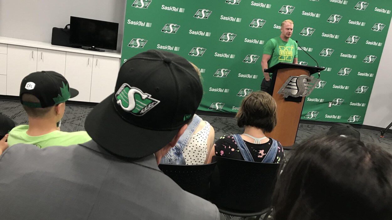 Jon Ryan devant un micro, devant le logo des Riders et devant un public venu assister à la conférence
