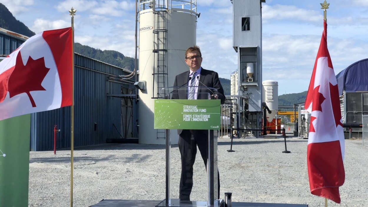 Un homme debout devant une tribune à l'extérieur d'une usine. Il est entouré de deux drapeaux canadiens.