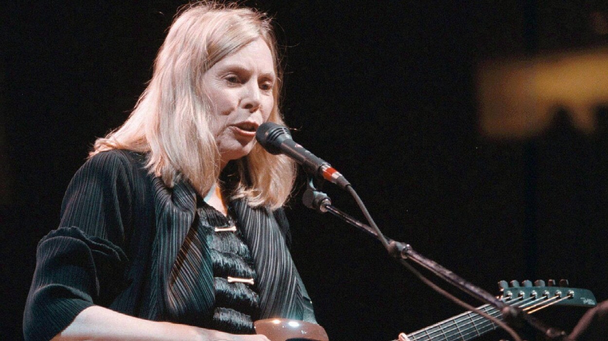 Joni Mitchell chante avec une guitare sur scène. Joni Mitchell chante avec une guitare sur scène.