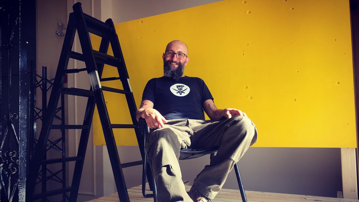 L'artiste José Luis Torres assis au coeur de son oeuvre, constituée d'une échelle noire et d'un panneau de bois peint en jaune.