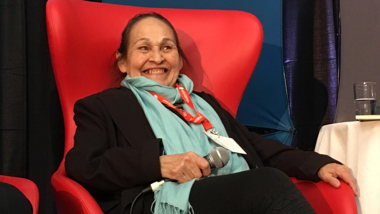 Joséphine Bacon, assise dans un fauteuil et tenant un micro, sourit