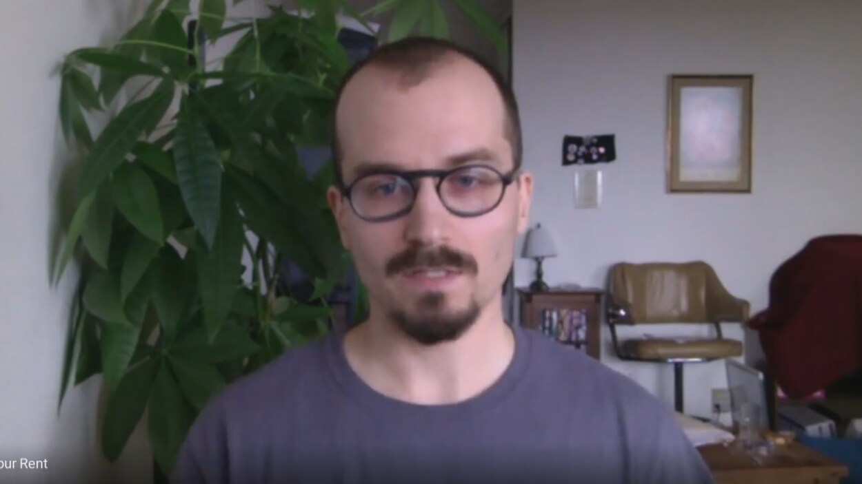 L'homme dans son salon, avec un grosse plante verte derrière lui.