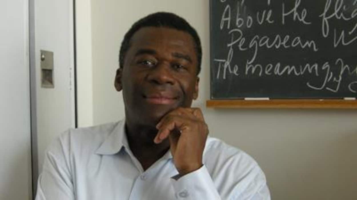 Portrait du poète d'origine haïtienne Joubert Satyre assis à un bureau, une main sous le menton, devant un tableau noir.
