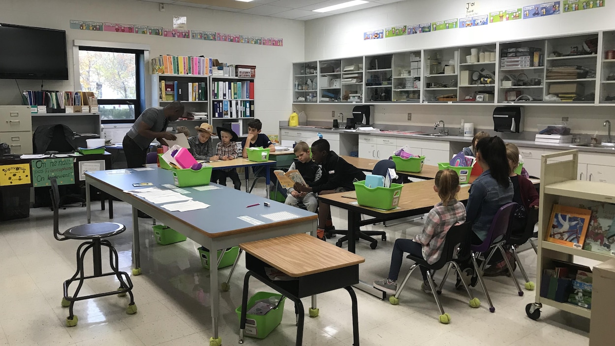 Une salle de classe avec quelques tables disposées en cercle. De jeunes enfants disposés en petits groupes et accompagnés de personnes plus âgées regardent des livres.