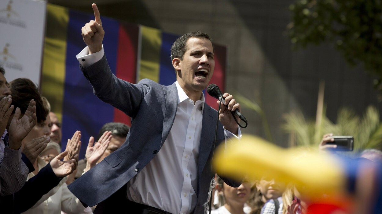 Juan Guaido parle dans un micro, sur une tribune. Des gens l'applaudissent.