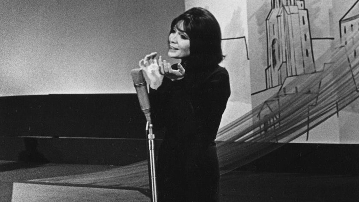 Juliette Greco chantant seule sur scène au micro dans une robe longue.