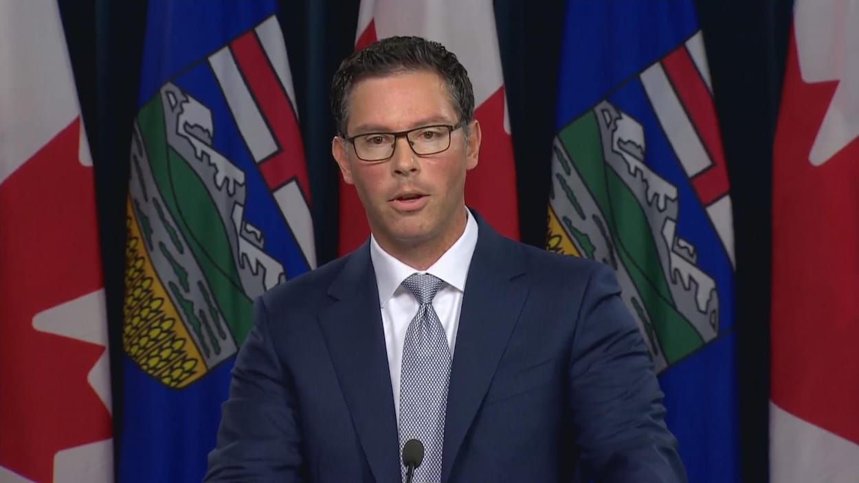 Un homme en costume cravate parle sur un podium devant une rangée de drapeaux de l'Alberta et du Canada.