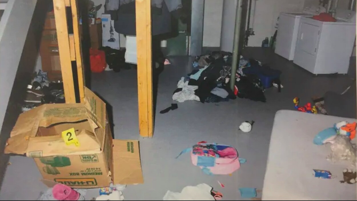 Un sous-sol sombre et encombré de divers objets, dont une boîte de carton vide et un tas de vêtements.
