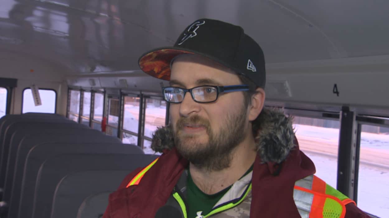 Justin Bucklashuk est de profil dans son autobus scolaire. Il est barbu et il porte une casquette, des lunettes et une veste de sécurité.