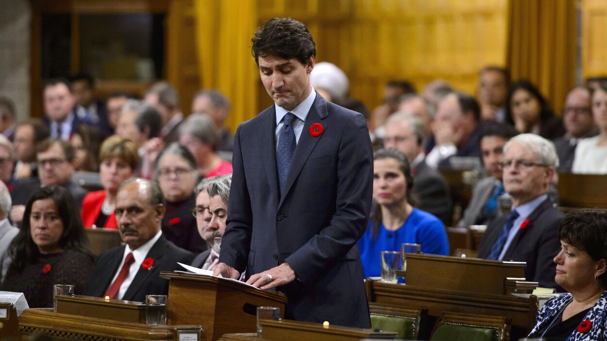 Justin Trudeau, debout parmi ses députés à la Chambre des communes, les yeux baissés et la mine contrite.