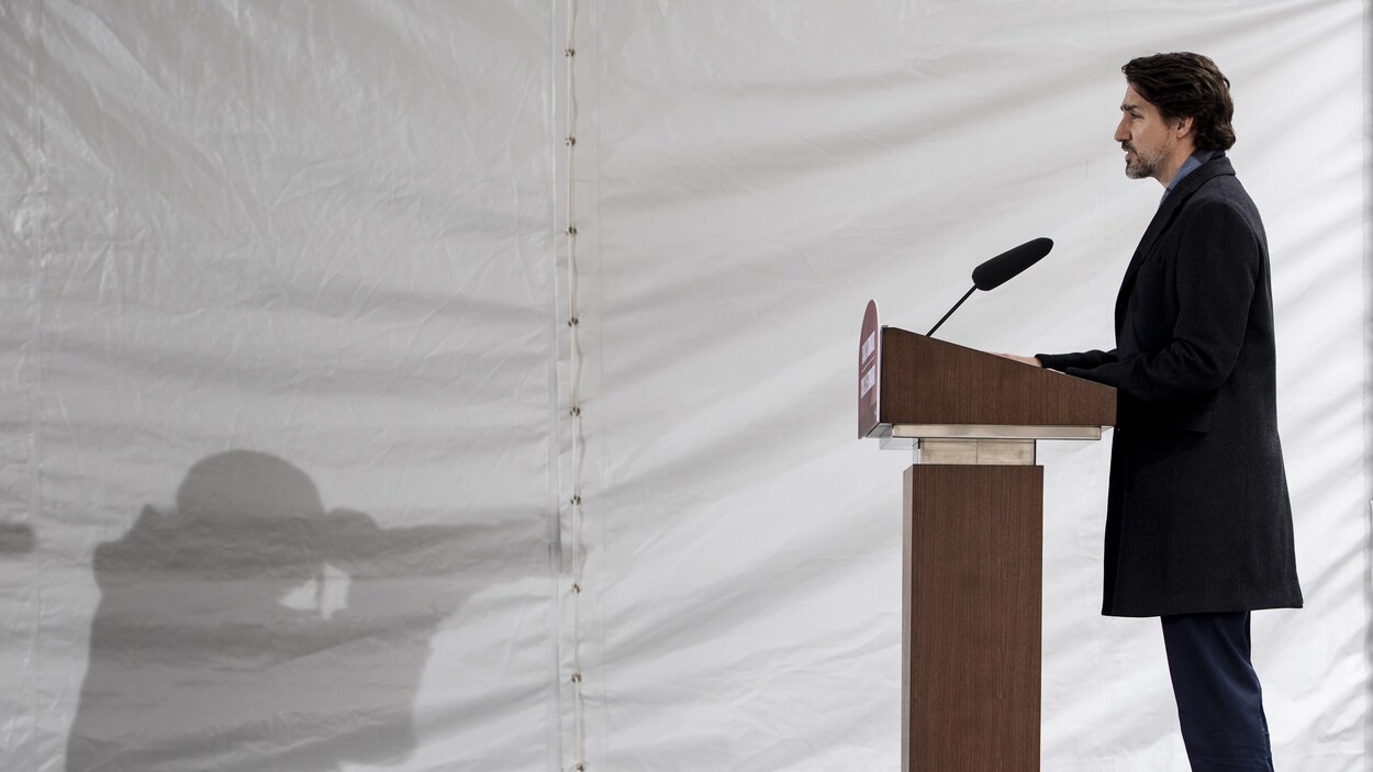 Justin Trudeau au lutrin lors d'une conférence de presse, avec l'ombre d'un photographe qui se reflète sur la toile blanche en arrière-plan.