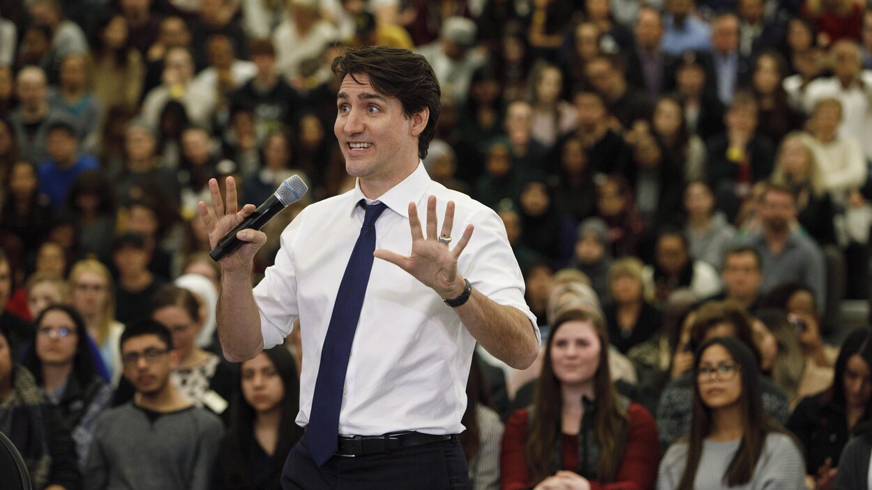 Justin Trudeau qui écarte les mains, tenant un micro devant une assemblée assise dans des gradins.