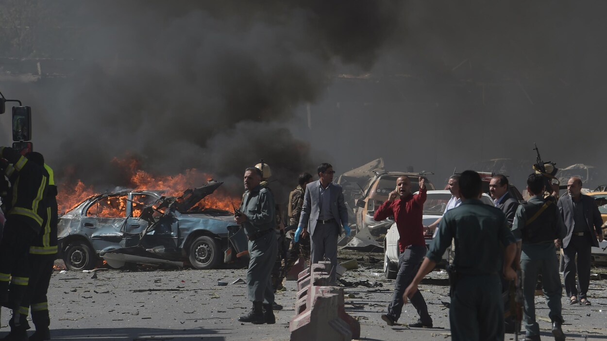 Plusieurs personnes dont des militaires et des secouristes sont sur les lieux de l'attentat, où une voiture en flamme laisse échapper beaucoup de fumée. Plusieurs personnes dont des militaires et des secouristes sont sur les lieux de l'attentat, où une voiture en flamme laisse échapper beaucoup de fumée.