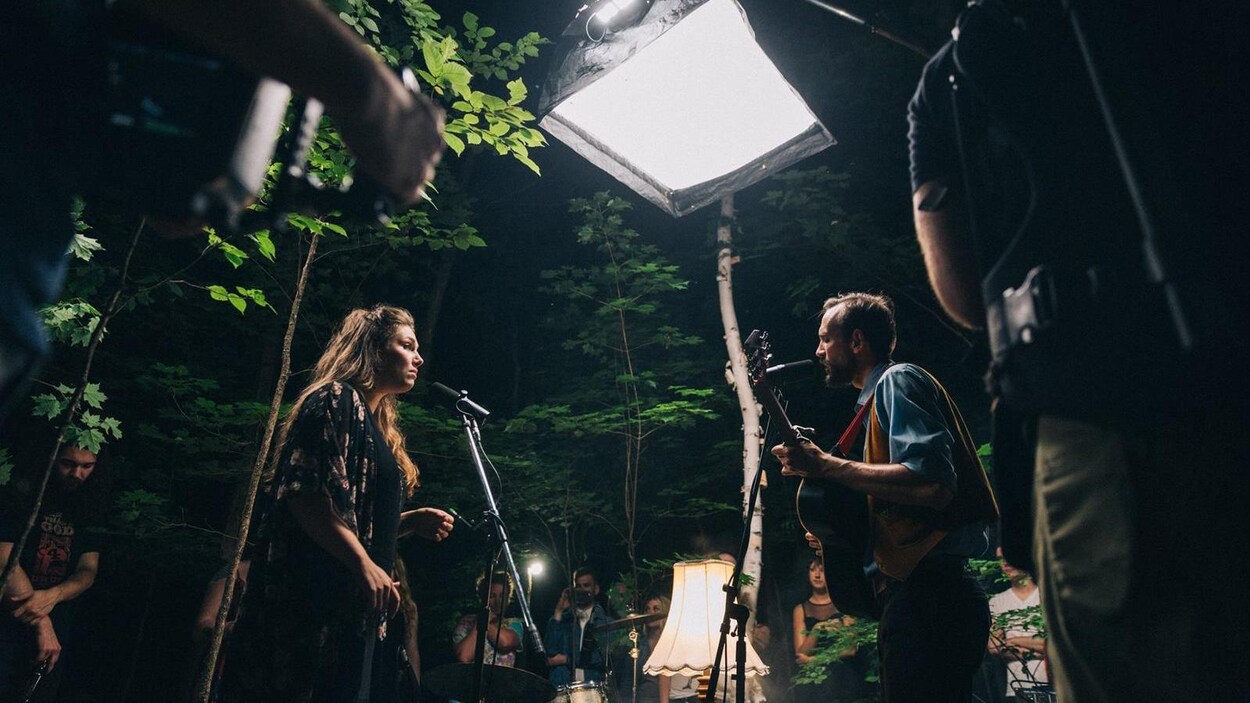 Une femme chante et un homme joue de la guitare sous un éclairage de tournage, entourés d'arbres et de quelques personnes. Une femme chante et un homme joue de la guitare sous un éclairage de tournage, entourés d'arbres et de quelques personnes.