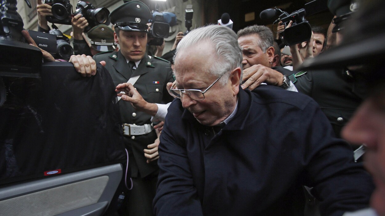 Fernando Karadima, qui porte des lunettes, s’apprête à monter dans une voiture, escorté par des policiers qui sont entourés de photographes.