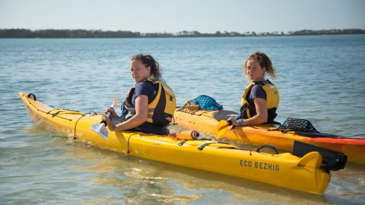 Deux jeunes femmes avec des cheveux frisés retenus par un élastique sont assises chacune dans un kayak, aviron à la main, sur l'eau et regardent en souriant l'objectif. Deux jeunes femmes avec des cheveux frisés retenus par un élastique sont assises chacune dans un kayak, aviron à la main, sur l'eau et regardent en souriant l'objectif.