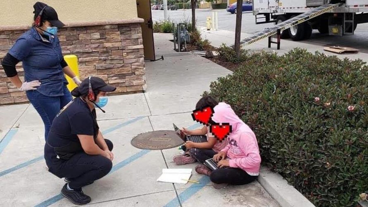 Deux filles penchées sur de petits ordinateurs portables, assises en tailleur sur le parterre en béton au bord d'un restaurant de la chaîne Taco Bell. Deux membres du personnel du restaurant s'adressent à elles.