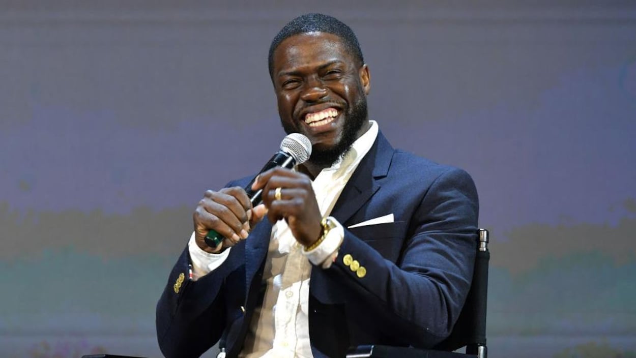 Kevin Hart, assis sur une chaise, tient un micro et rit.