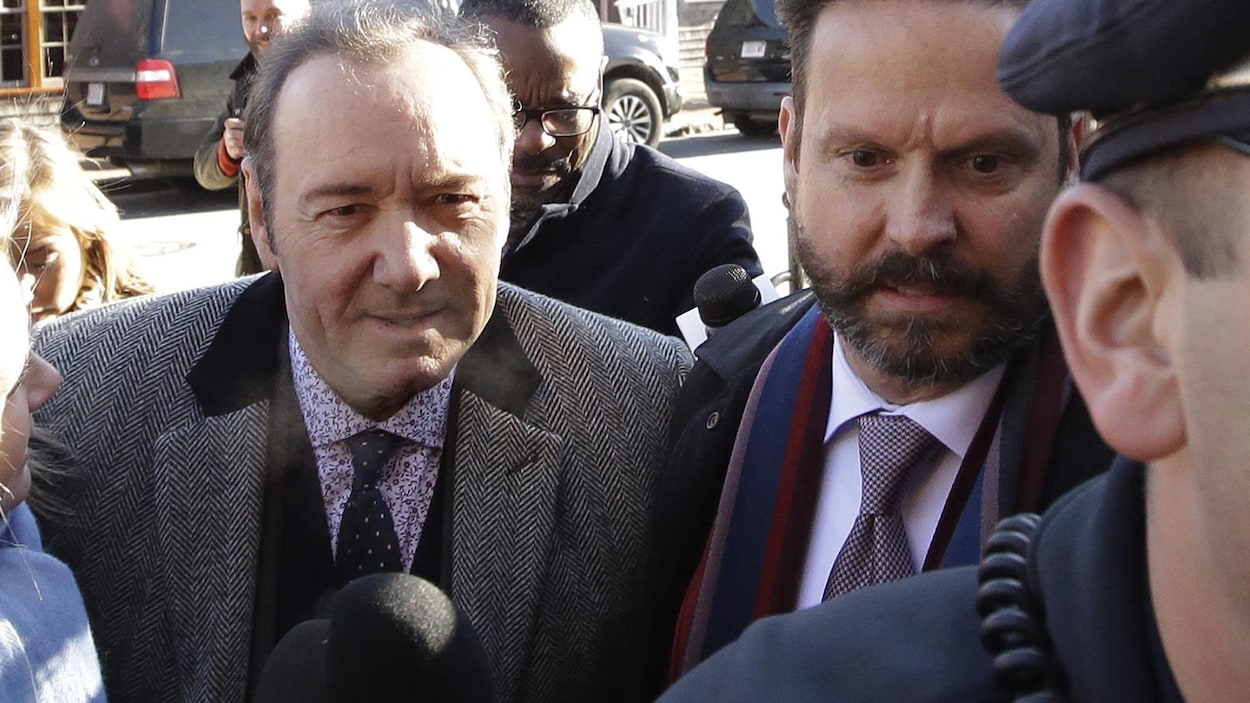 L'homme est entouré de journalistes avec des micros.