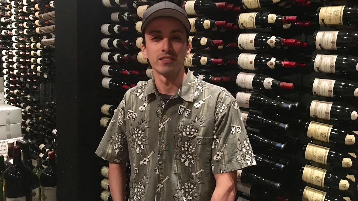 Un homme à casquette posant devant des étagères de bouteilles de vins.