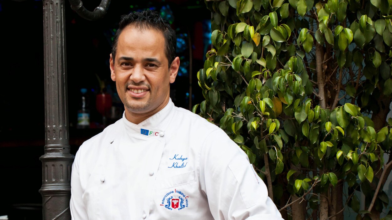 Khalid Kahya, vêtu de son uniforme de chef, accoté sur un lampadaire, devant un arbre.