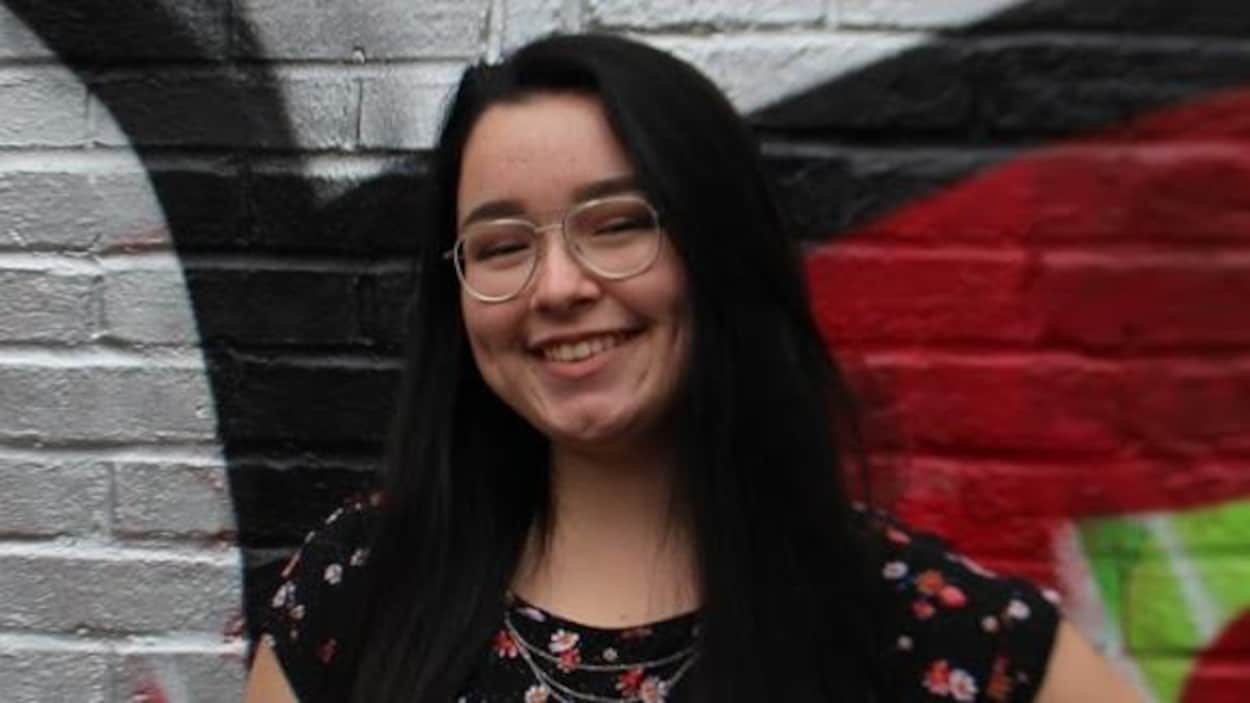 Une jeune femme sourit à la caméra devant un mur graffité.