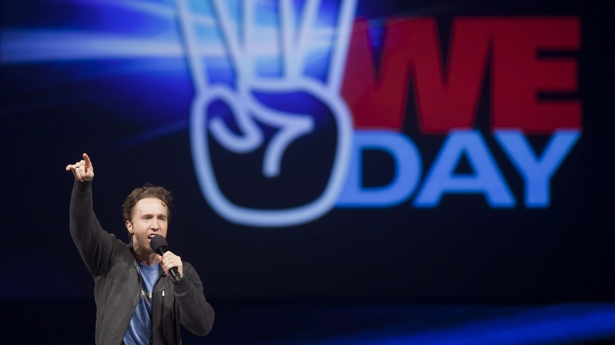 Un homme parle dans un micro sur une scène avec une affiche « We Day » en arrière-plan.